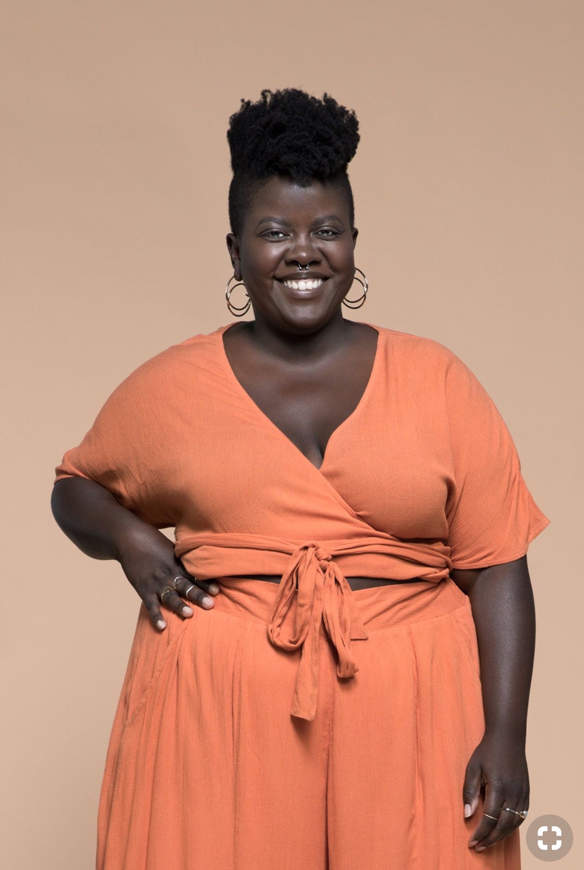 A woman is smiling and wearing a coral wrap top and pants 